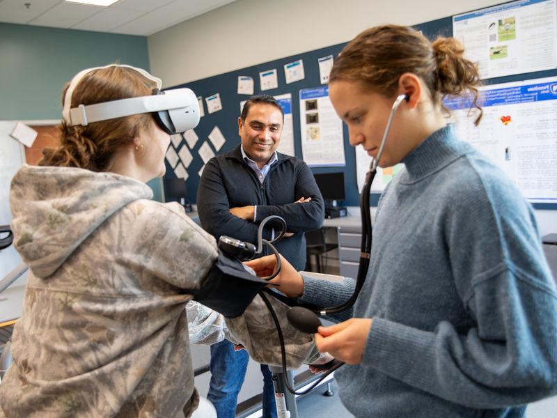 Student takes blood pressure of another student on stationary bike wearing VR goggles while faculty observes